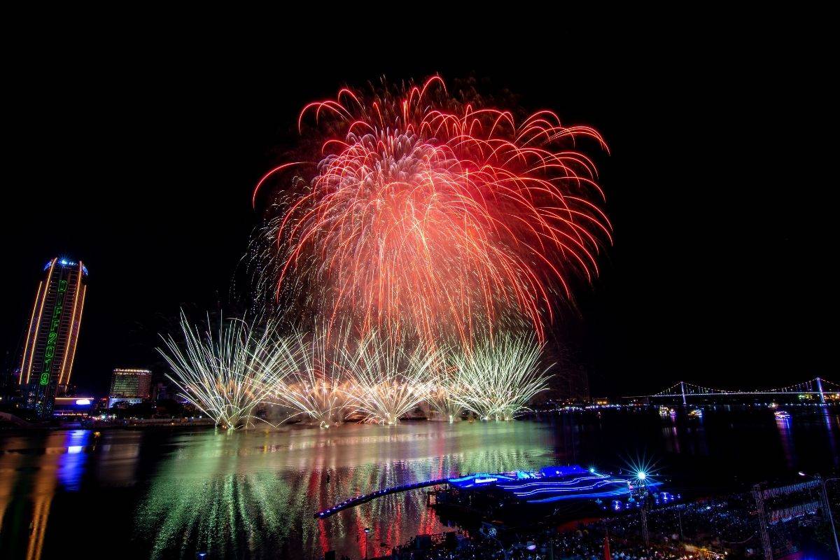 fireworks exploding over a river with city lights reflecting on the water