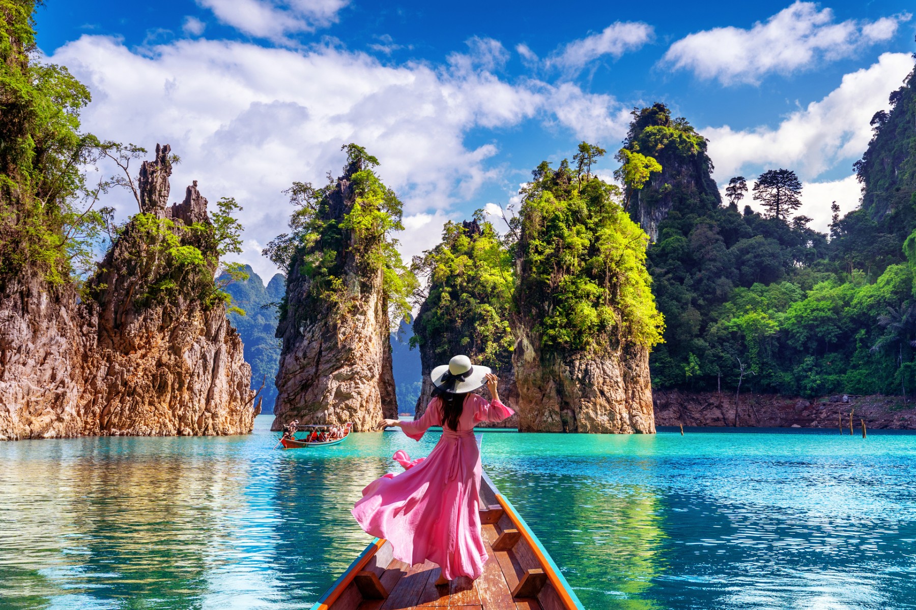 Woman in pink dress standing on a longtail boat, admiring the limestone cliffs at Ratchaprapha Dam in Khao Sok National Park, Thailand