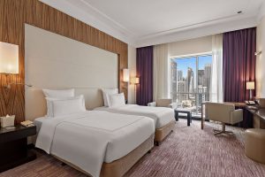 Modern twin bedroom at Pullman Dubai Jumeirah Lakes Towers featuring two single beds, stylish wood paneling, and a large window view.