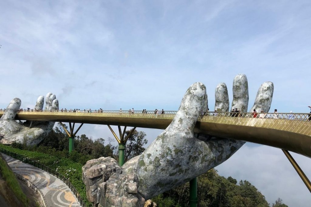 tall golden bridge held up by two giant stone hands above green hills and mist